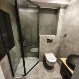 Nowoczesna łazienka z kabiną prysznicową z czarnymi profilami, podwieszaną toaletą i umywalką nablatową na drewnianym blacie, wykończona szarymi płytkami imitującymi beton.