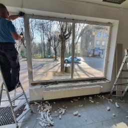 EPP Oskar Kasperkowiak - Pracownik na drabinie montuje okno w lokalu, widoczne narzędzia, resztki pianki montażowej na podłodze i widok na ulicę z niebieskim samochodem.