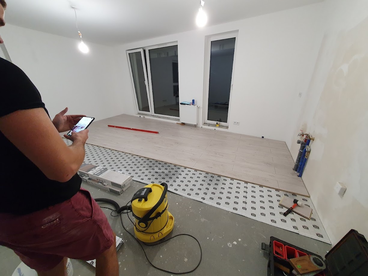 W trakcie montażu jasnych paneli podłogowych w pustym pokoju, widoczna folia izolacyjna, poziomica, odkurzacz przemysłowy i osoba robiąca zdjęcie telefonem.