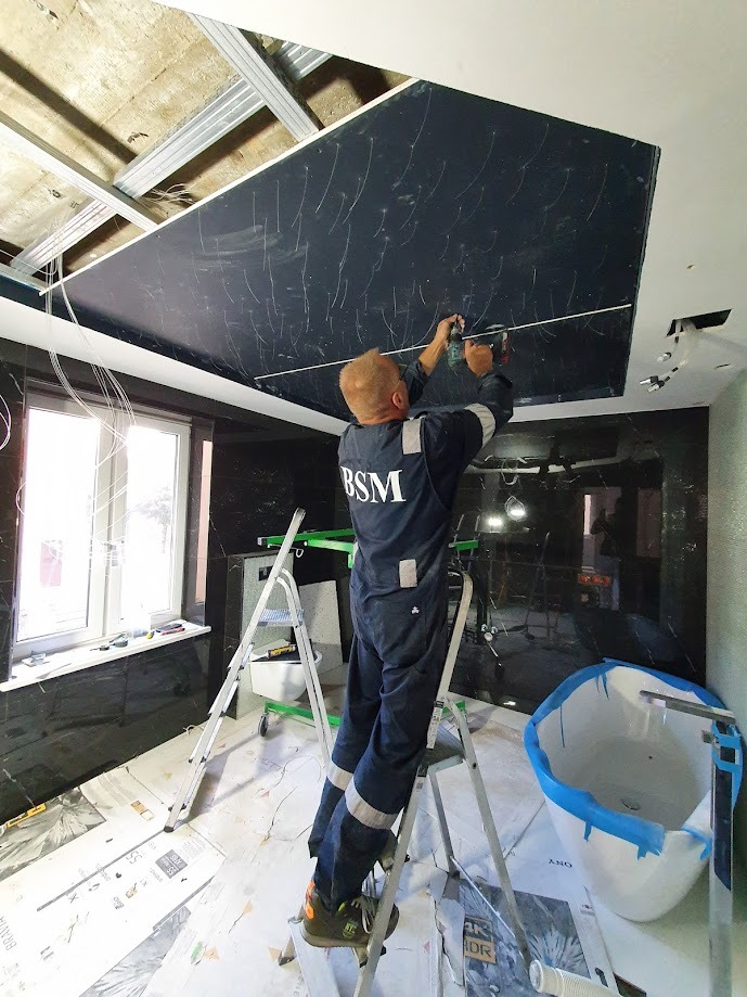 Pracownik w kombinezonie BSM montuje czarny panel sufitowy z wzorem przypominającym marmur, stojąc na aluminiowej drabinie w łazience z marmurowymi ścianami, widoczne metalowe profile konstrukcyjne...