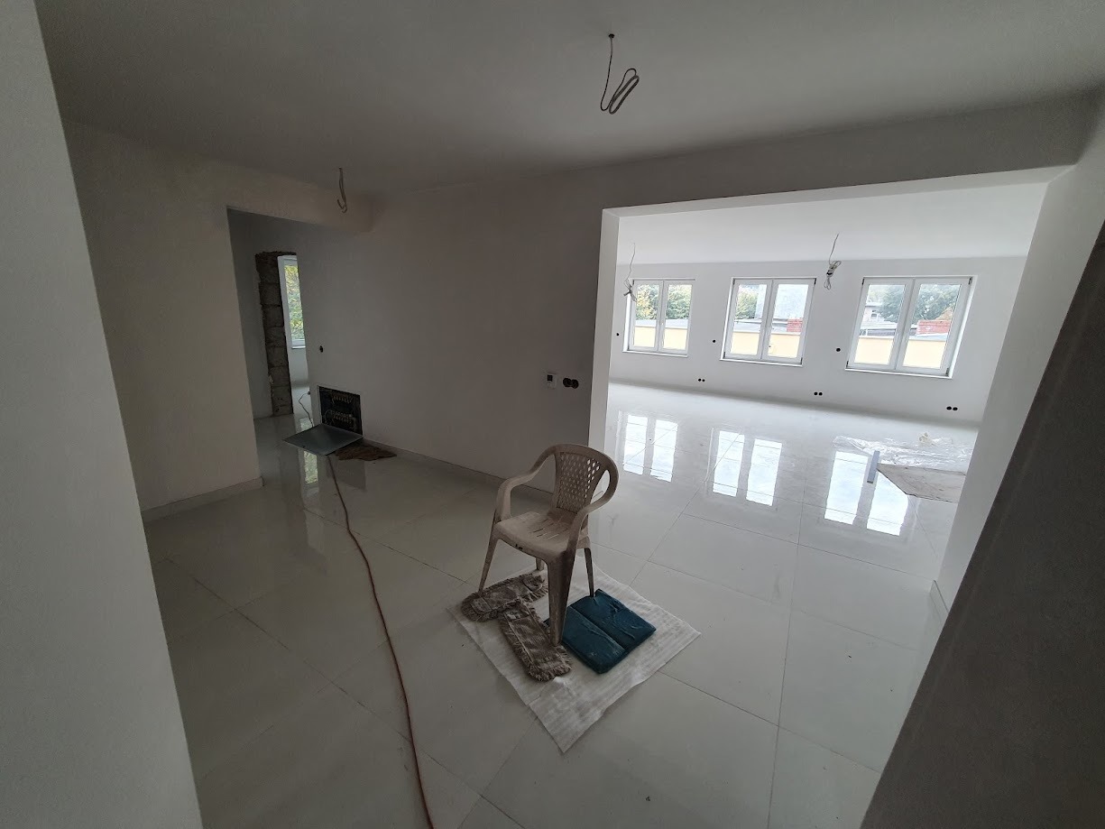 Wnętrze w stanie deweloperskim z białymi ścianami i błyszczącą, jasną podłogą, plastikowe krzesło i narzędzia malarskie na środku pomieszczenia, widoczne kable wystające z sufitu.