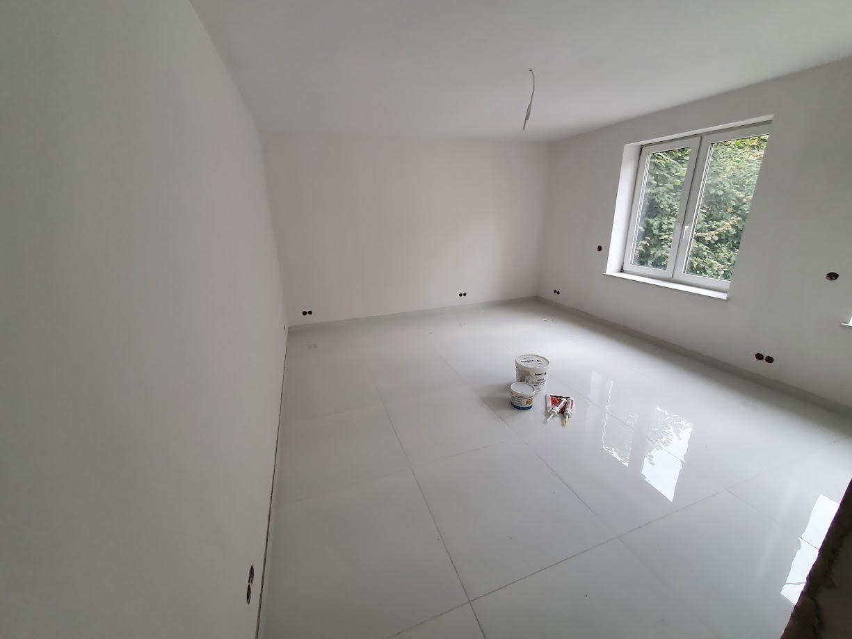 Pomalowane na biało puste pomieszczenie z błyszczącą podłogą, widoczne puszki z farbą i tubki z masą uszczelniającą, okno z widokiem na zieleń.