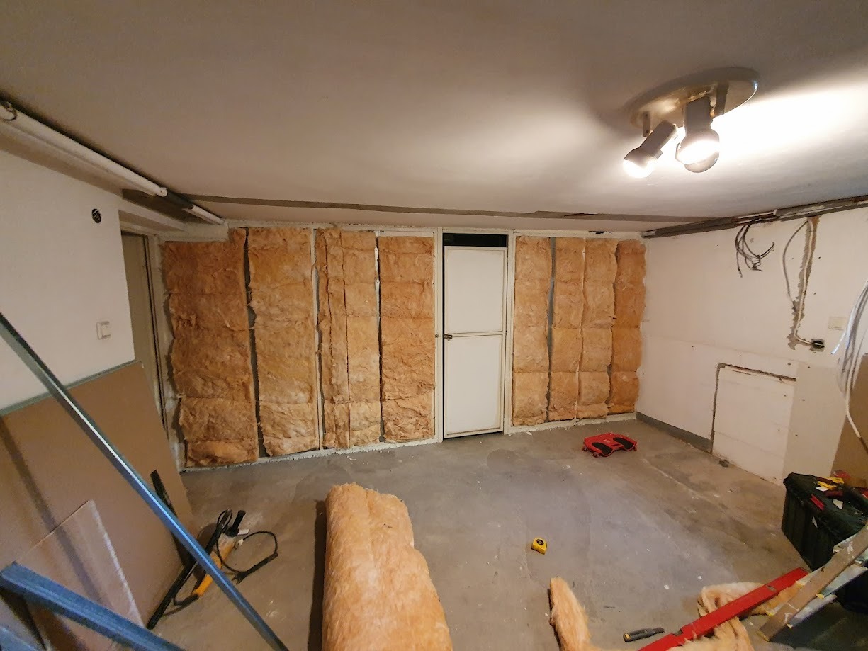 Wnętrze w trakcie prac izolacyjnych ścian wełną mineralną w metalowym stelażu, widoczne narzędzia i materiały budowlane na podłodze, surowe wykończenie.