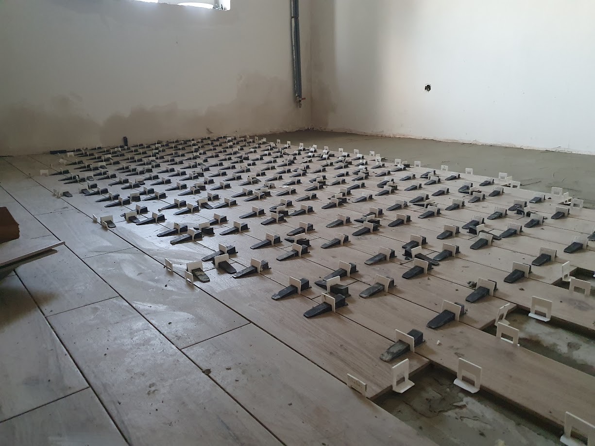 Ułożone płytki podłogowe z systemem poziomowania, widoczne klipsy i kliny, surowy stan ścian w tle.