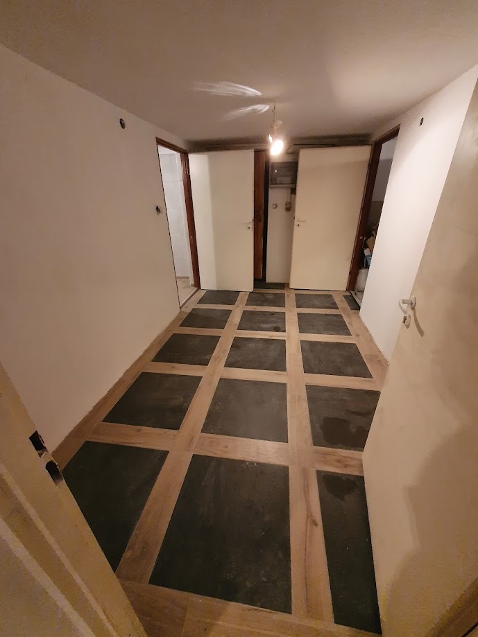 Korytarz z nową podłogą w kwadraty: ciemne płyty otoczone jasnym drewnem, białe ściany i drzwi, surowy klimat, widoczne punkty świetlne na suficie.