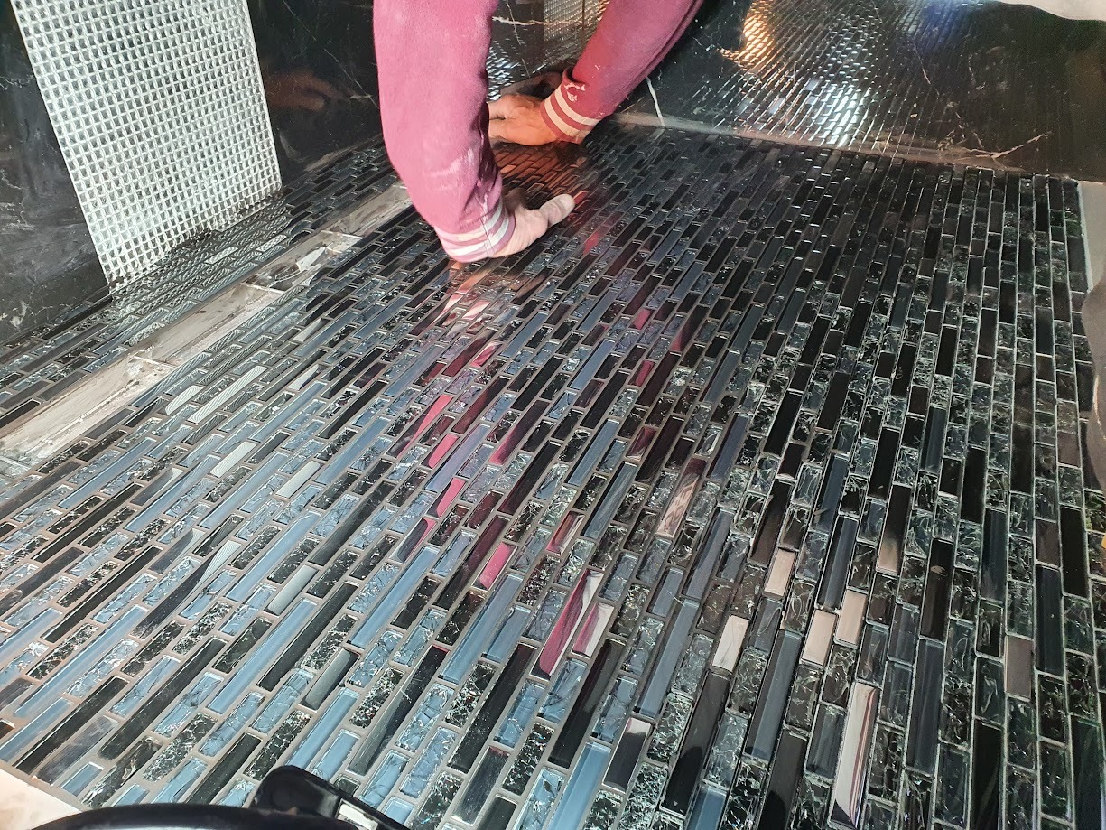 Układanie mozaiki szklanej w odcieniach szarości i czerni na ścianie, widoczne ręce wykonawcy w różowej bluzie z białymi mankietami.