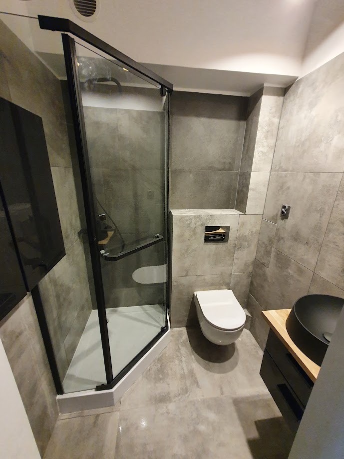 Nowoczesna łazienka z kabiną prysznicową z czarnymi profilami, podwieszaną toaletą i umywalką nablatową na drewnianym blacie, wykończona szarymi płytkami imitującymi beton.