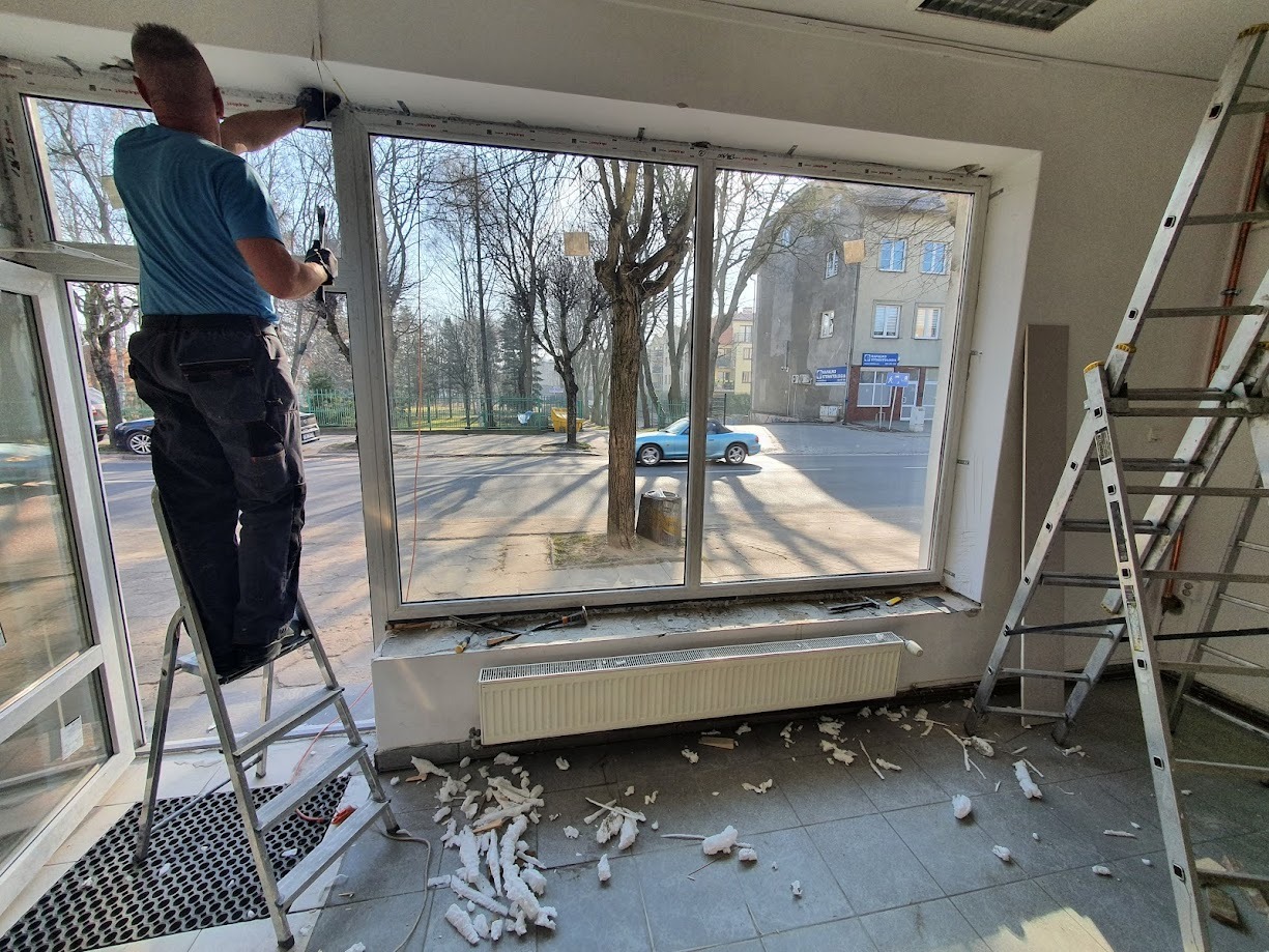 Pracownik na drabinie montuje okno w lokalu, widoczne narzędzia, resztki pianki montażowej na podłodze i widok na ulicę z niebieskim samochodem.