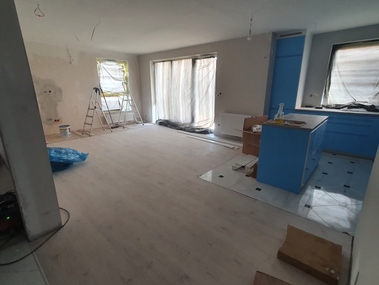 Wnętrze mieszkania w trakcie remontu: położone jasne panele podłogowe, ściany przygotowane do malowania, okna zabezpieczone folią, widoczna drabina malarska i fragment niebieskiej zabudowy kuchennej.