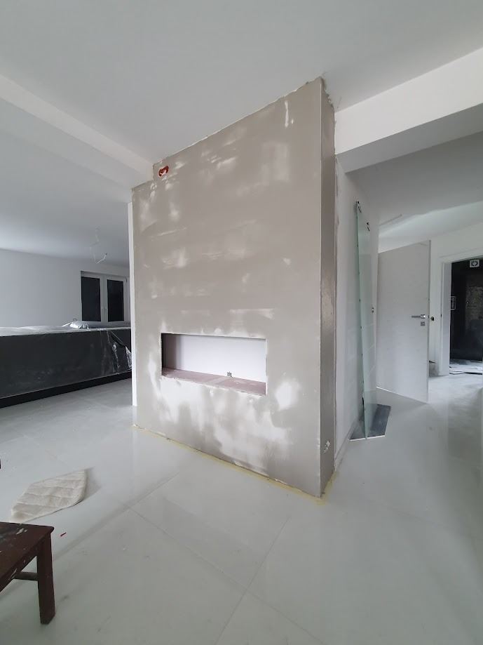 Ściana działowa w trakcie prac wykończeniowych, z widocznym otworem wnęki, pokryta szpachlą, z widocznymi śladami po narzędziach i niedoskonałościami powierzchni, w jasnym, minimalistycznym wnętrzu...