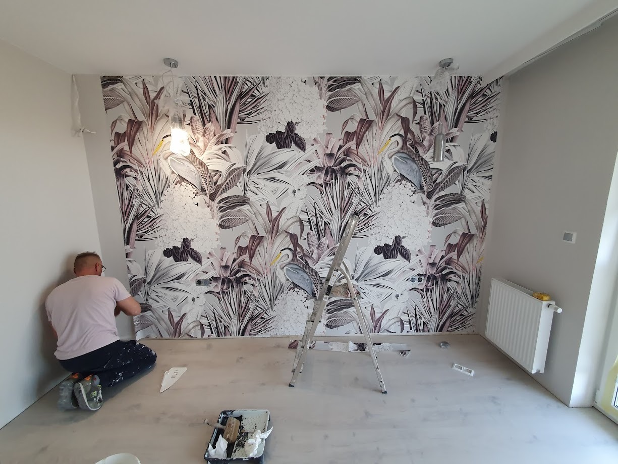 Mężczyzna w trakcie tapetowania ściany wzorzystą tapetą w motywy roślinne i ptasie, widoczna drabina aluminiowa, narzędzia malarskie i grzejnik.