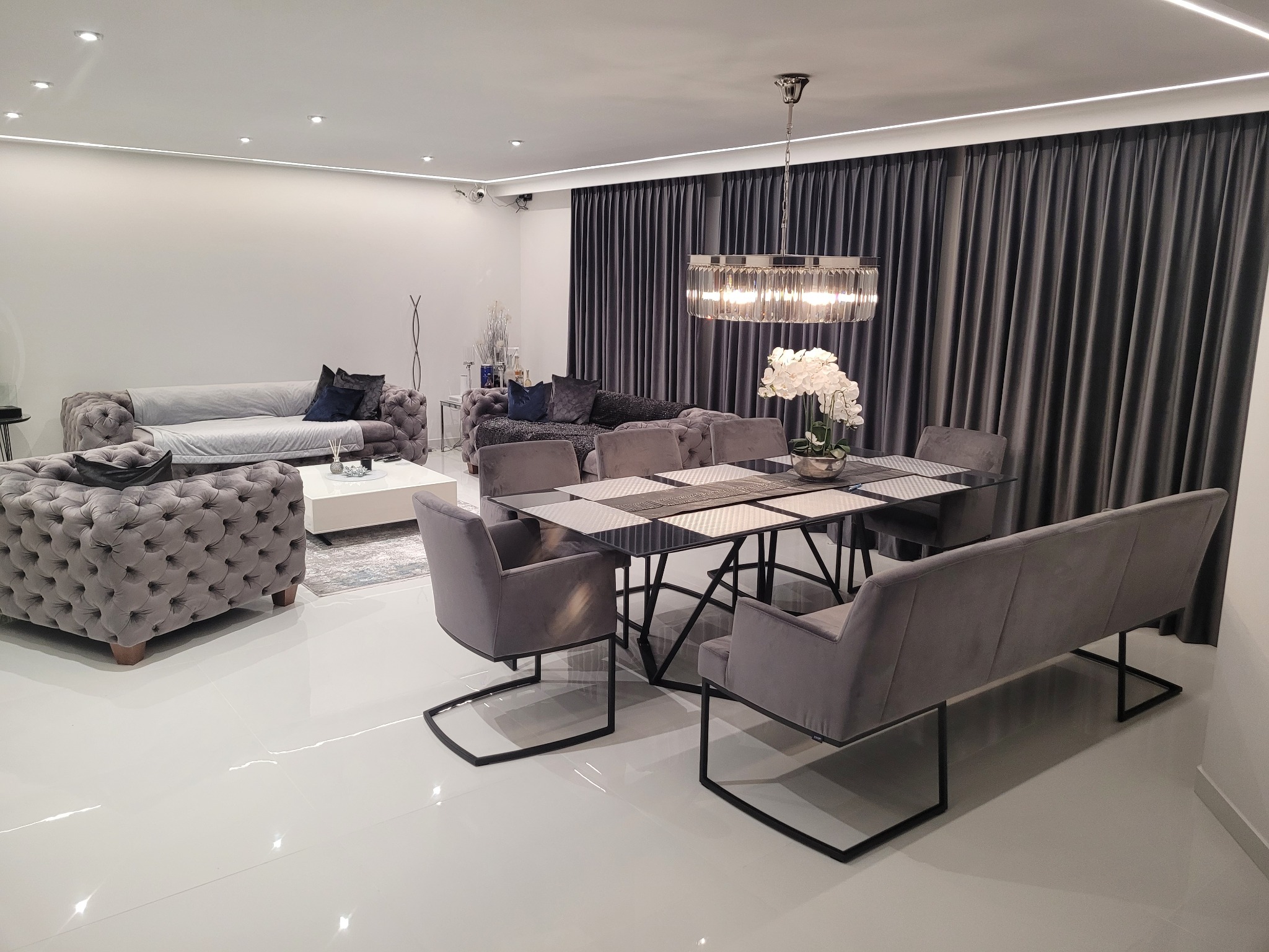Eleganckie wnętrze salonu z szarymi meblami tapicerowanymi, szklanym stołem jadalnym, kryształowym żyrandolem i białymi błyszczącymi płytkami podłogowymi, oświetleniem LED na suficie i zasłonami...