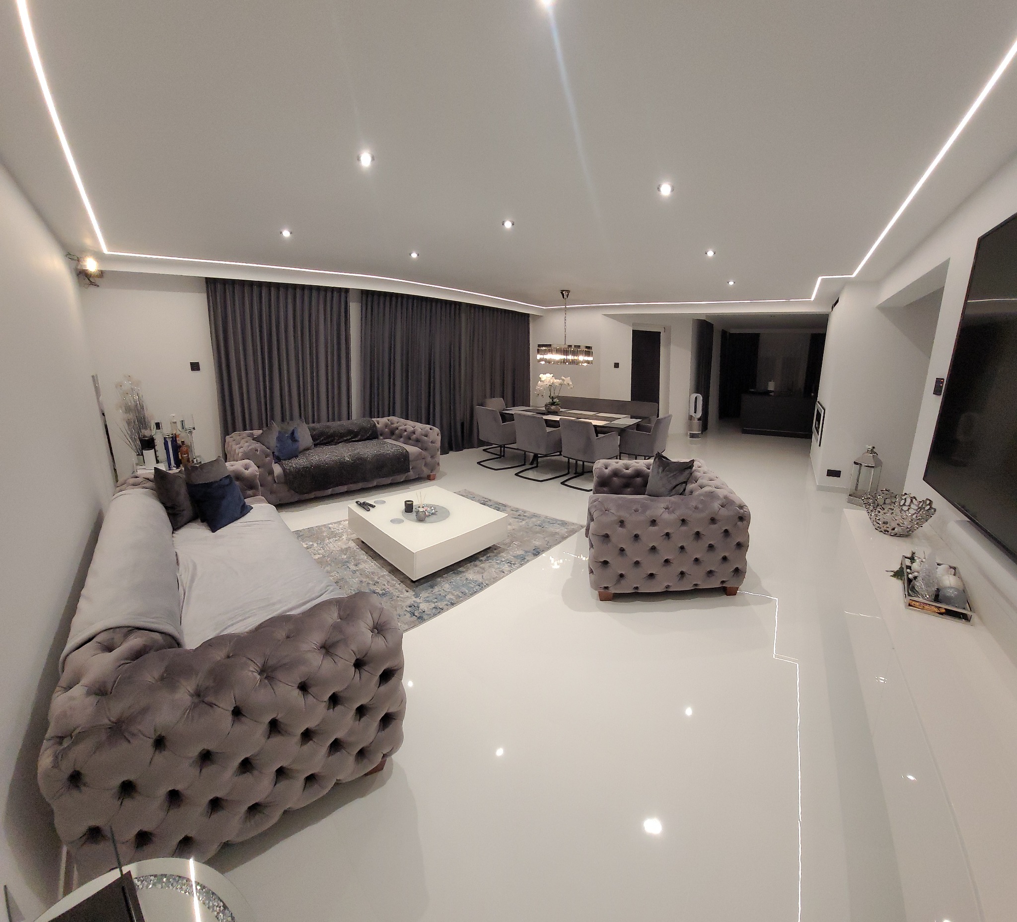 Przestronny salon z szarymi, pikowanymi sofami, białym stolikiem kawowym na wzorzystym dywanie i połyskującą białą podłogą, oświetlony listwami LED pod sufitem.