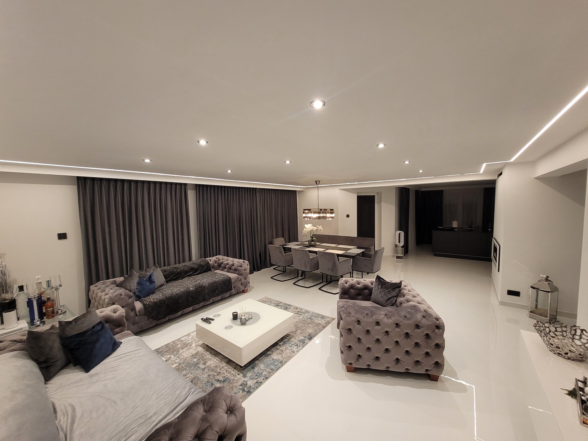 Przestronny salon z szarymi, pikowanymi sofami, białym stolikiem kawowym na wzorzystym dywanie, szarymi zasłonami i stołem z krzesłami w tle, oświetlony punktowymi lampami sufitowymi i listwami LED.