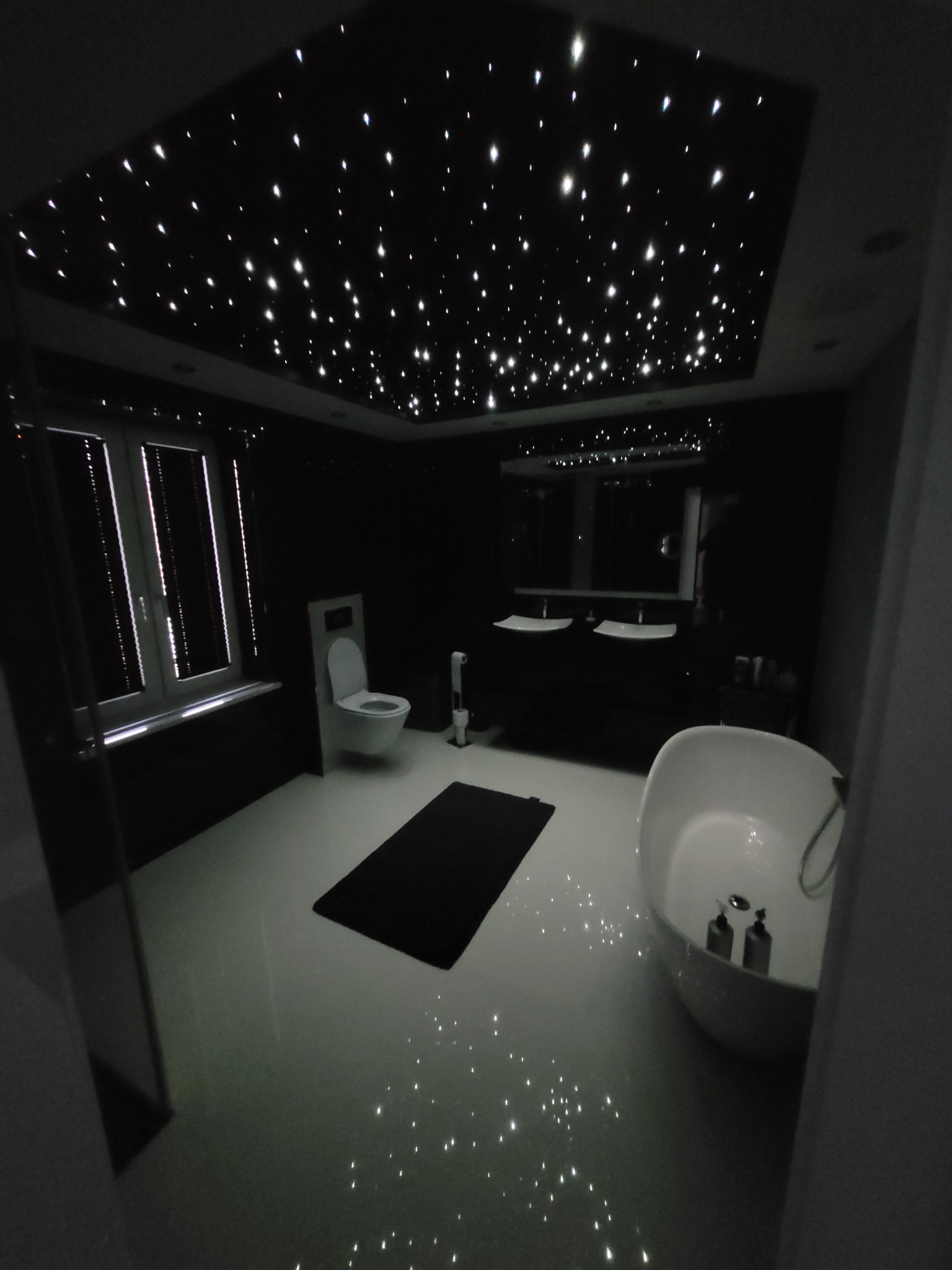 Nowoczesna łazienka w ciemnej tonacji z sufitem imitującym rozgwieżdżone niebo, owalna wanna wolnostojąca i podwójna umywalka na tle lustra.