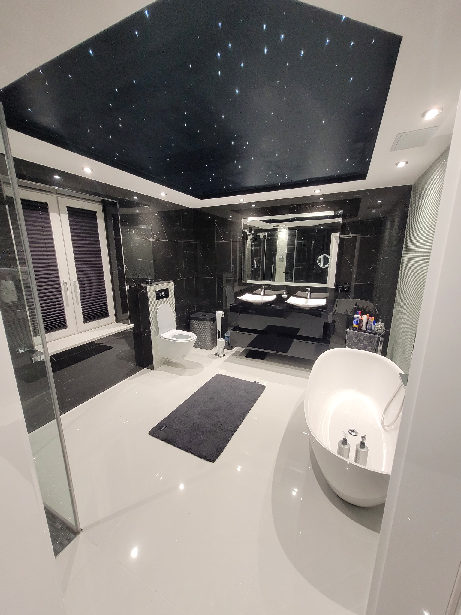 Nowoczesna łazienka z czarnymi marmurowymi ścianami, białymi płytkami podłogowymi i sufitem imitującym nocne niebo z gwiazdami, oświetlonym punktowymi lampami.