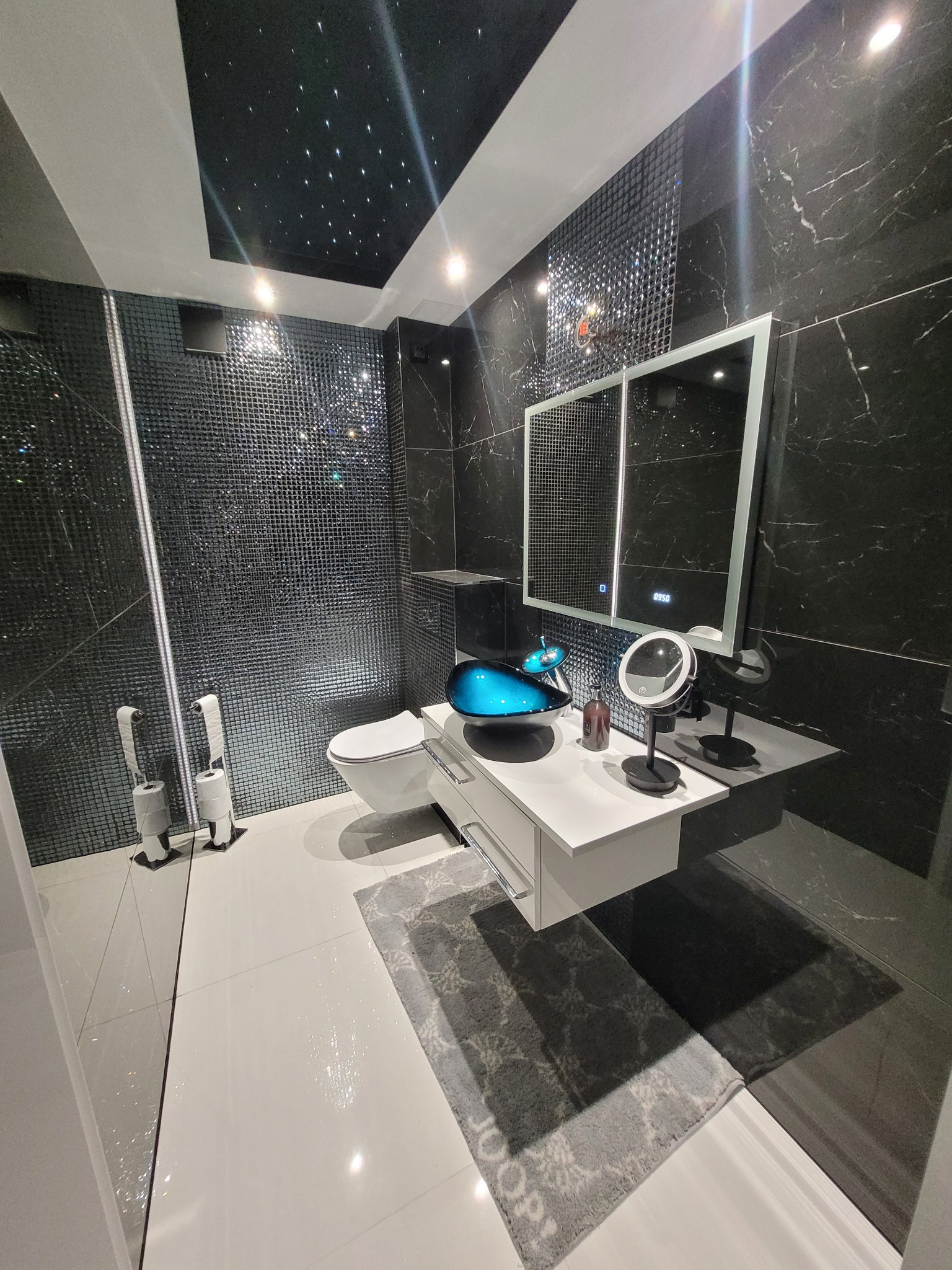 Nowoczesna łazienka z czarno-białymi płytkami marmurkowymi, podświetlanym sufitem imitującym gwiazdy, białą szafką pod umywalką z turkusową misą i szarą matą podłogową.