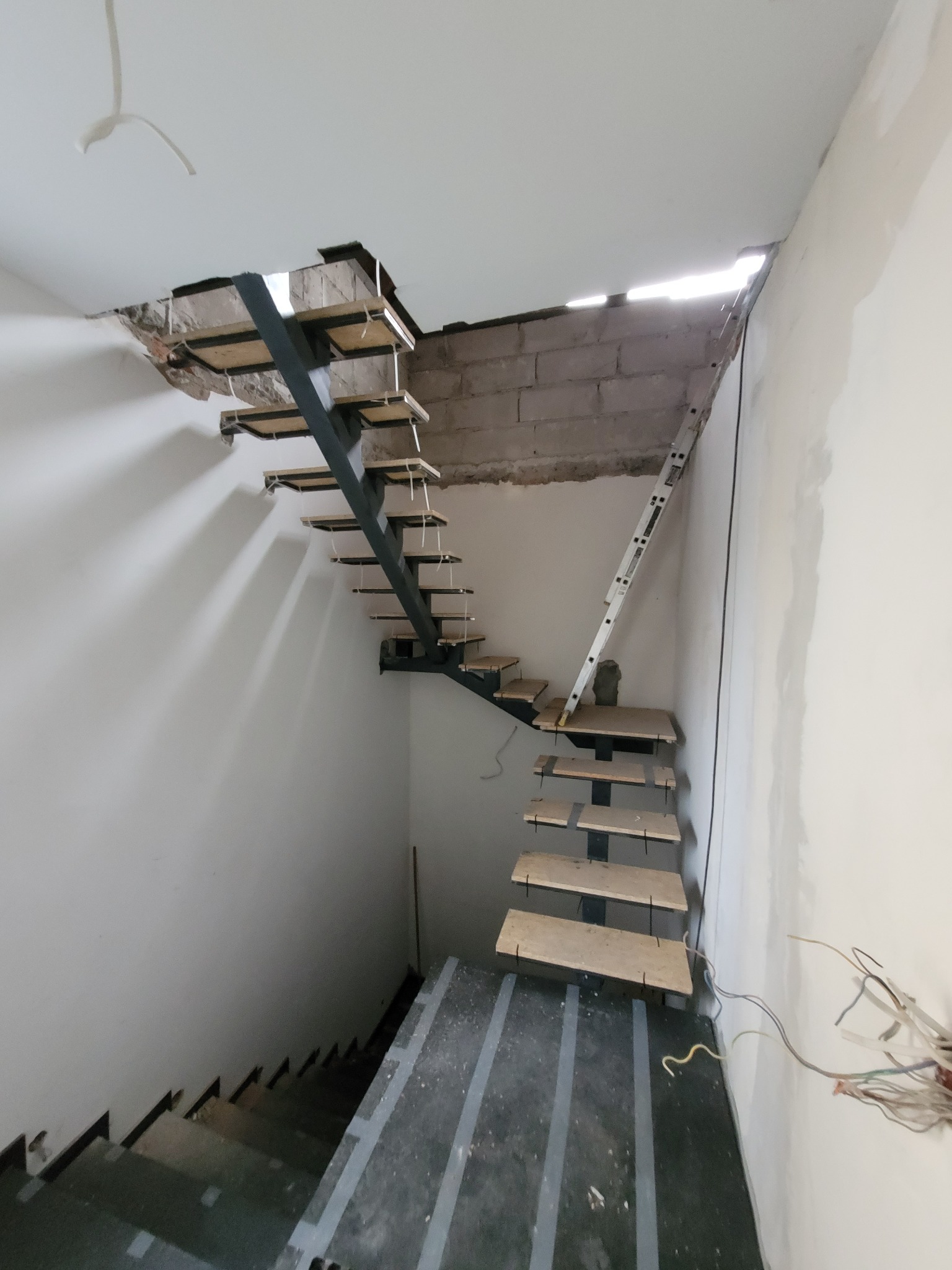 Montaż schodów ażurowych z metalową konstrukcją i drewnianymi stopniami w surowym wnętrzu, widoczne niedokończone ściany i otwór w suficie, drabina oparta o ścianę.