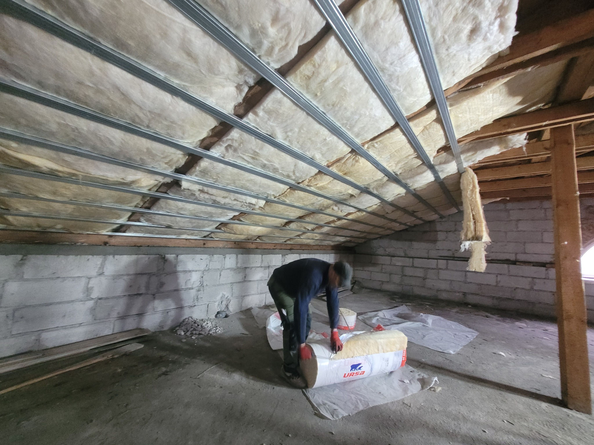 Pracownik w rękawicach tnie nożem rolkę wełny mineralnej URSA na poddaszu z częściowo zaizolowanym sufitem, widoczne metalowe profile montażowe.