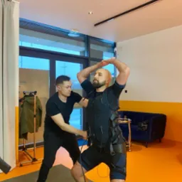 Trener personalny zakłada kamizelkę EMS na mężczyznę z brodą, który stoi w rozkroku w skarpetkach w kosmos.