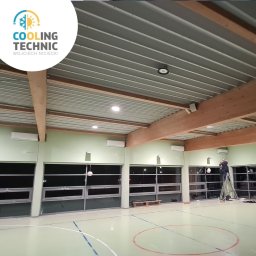 Cooling Technic Wojciech Niciecki - Montaż klimatyzacji w hali sportowej: widoczne jednostki wewnętrzne pod sufitem, pracownik na drabinie podczas instalacji, oświetlenie halogenowe, sufit z blachy trapezowej.