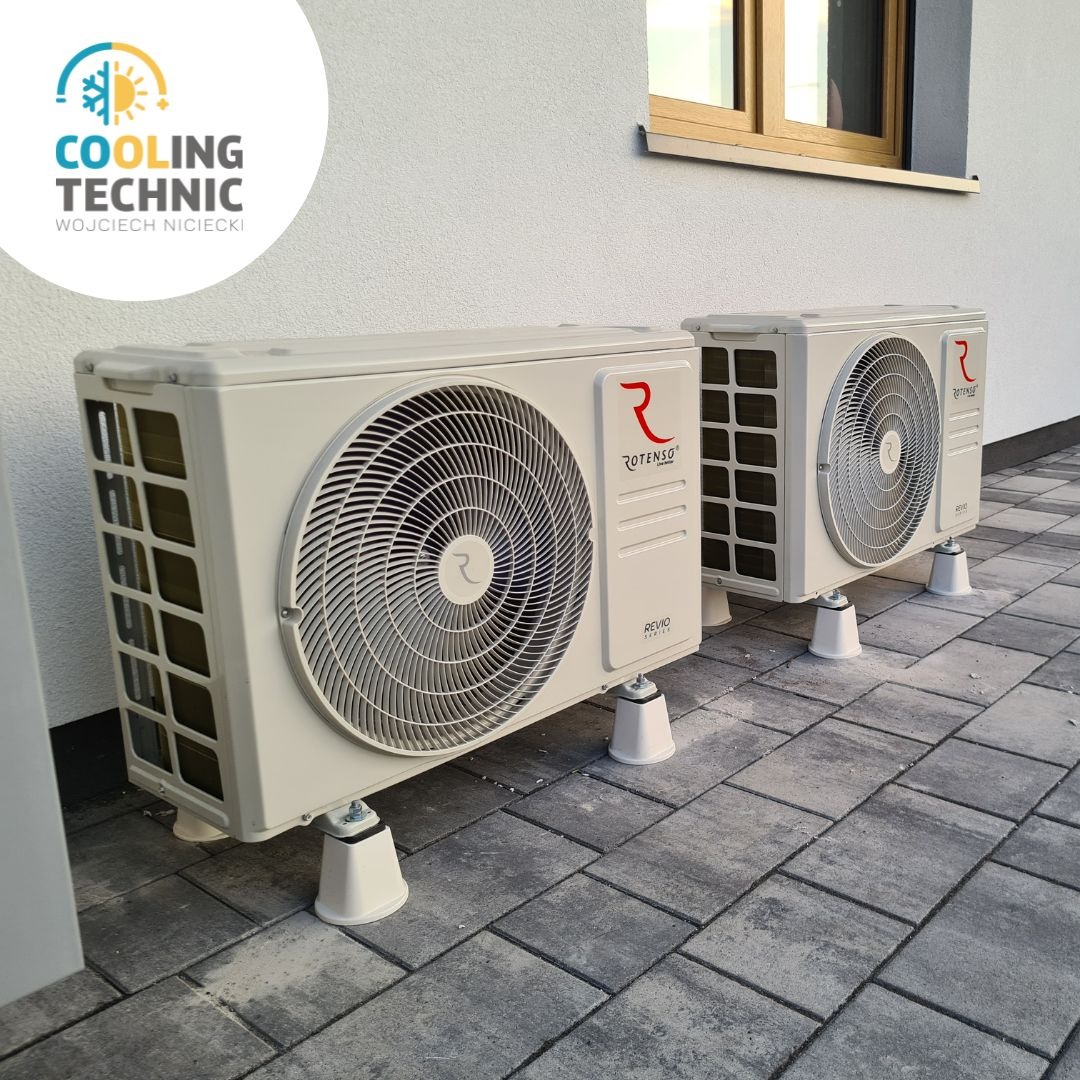 Dwa identyczne, beżowe agregaty klimatyzacji marki Rotenso, model Revio Series, zamontowane na białych podstawkach na zewnątrz budynku z szarą kostką brukową i oknem w tle.