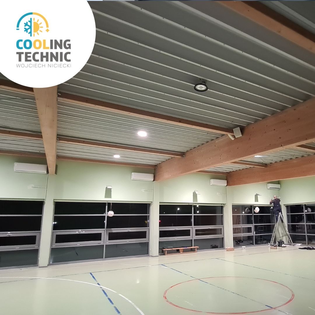 Montaż klimatyzacji w hali sportowej: widoczne jednostki wewnętrzne pod sufitem, pracownik na drabinie podczas instalacji, oświetlenie halogenowe, sufit z blachy trapezowej.