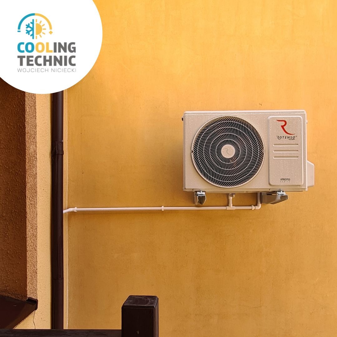 Beżowy klimatyzator Rotenso zamontowany na żółtej ścianie budynku, widoczne rury instalacyjne.