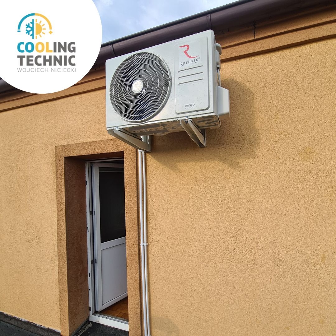 Zamontowana jednostka zewnętrzna klimatyzatora Rotenso na ścianie budynku z beżową elewacją, widoczna brama wejściowa i rynna dachowa.