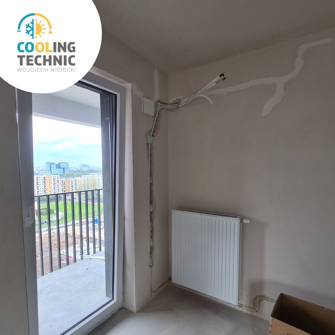 Wnętrze w trakcie prac instalacyjnych: przewody elektryczne prowadzone po ścianie, częściowo zabezpieczone pianką montażową, widoczny grzejnik i wyjście na balkon z widokiem na miasto.