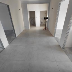 Usługi remontowo budowlane - Ułożona szara podłoga z płytek w korytarzu, widoczne przygotowane ościeżnice drzwiowe i fragment wykończonej łazienki w głębi. Na podłodze stoi lampa budowlana.