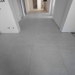 Usługi remontowo budowlane - Jasnoszara, duża płytka podłogowa w korytarzu z białymi ścianami i otworami drzwiowymi. W tle widoczne pomieszczenia w trakcie wykańczania.