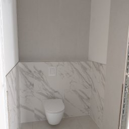 Usługi remontowo budowlane - Wykończona łazienka z toaletą wiszącą i płytkami imitującymi marmur na ścianach do połowy wysokości. Górna część ścian wyłożona szarymi, podłużnymi płytkami.