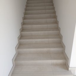 Usługi remontowo budowlane - Wykończone schody wewnętrzne z jasnych płytek ceramicznych z delikatnym wzorem marmuru, z antypoślizgowymi paskami na krawędziach stopni, przy białej ścianie.