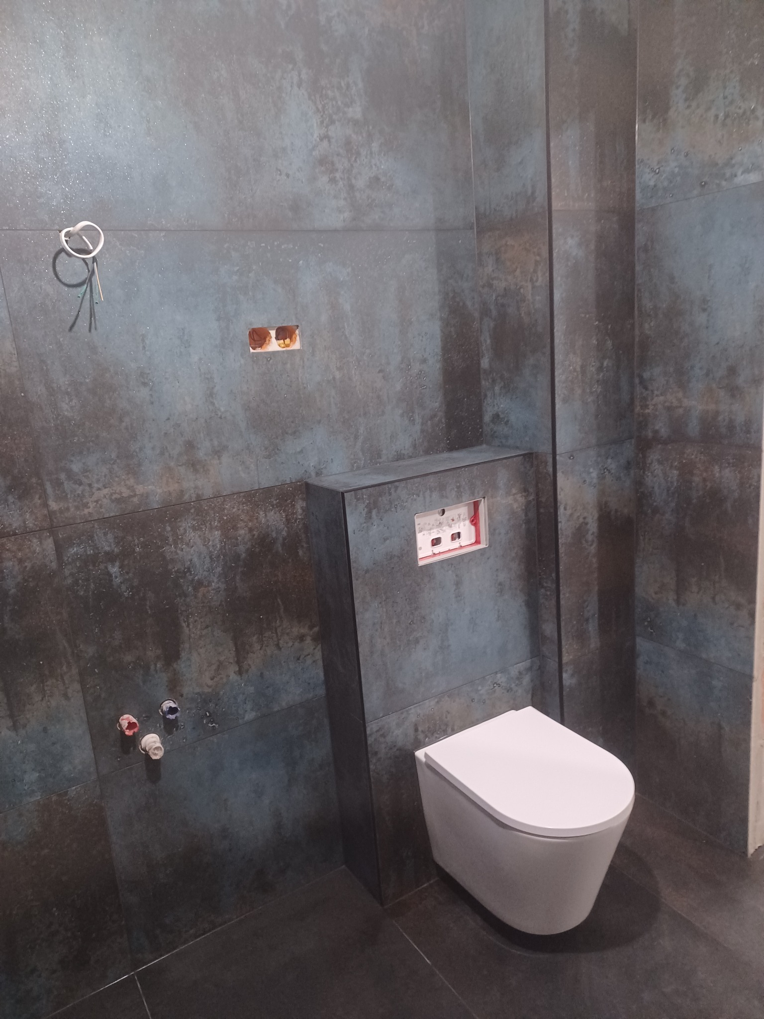 Podwieszana toaleta z zabudowaną spłuczką na tle ściany wyłożonej płytkami imitującymi rdzę, widoczne wystające kable i rury instalacyjne.