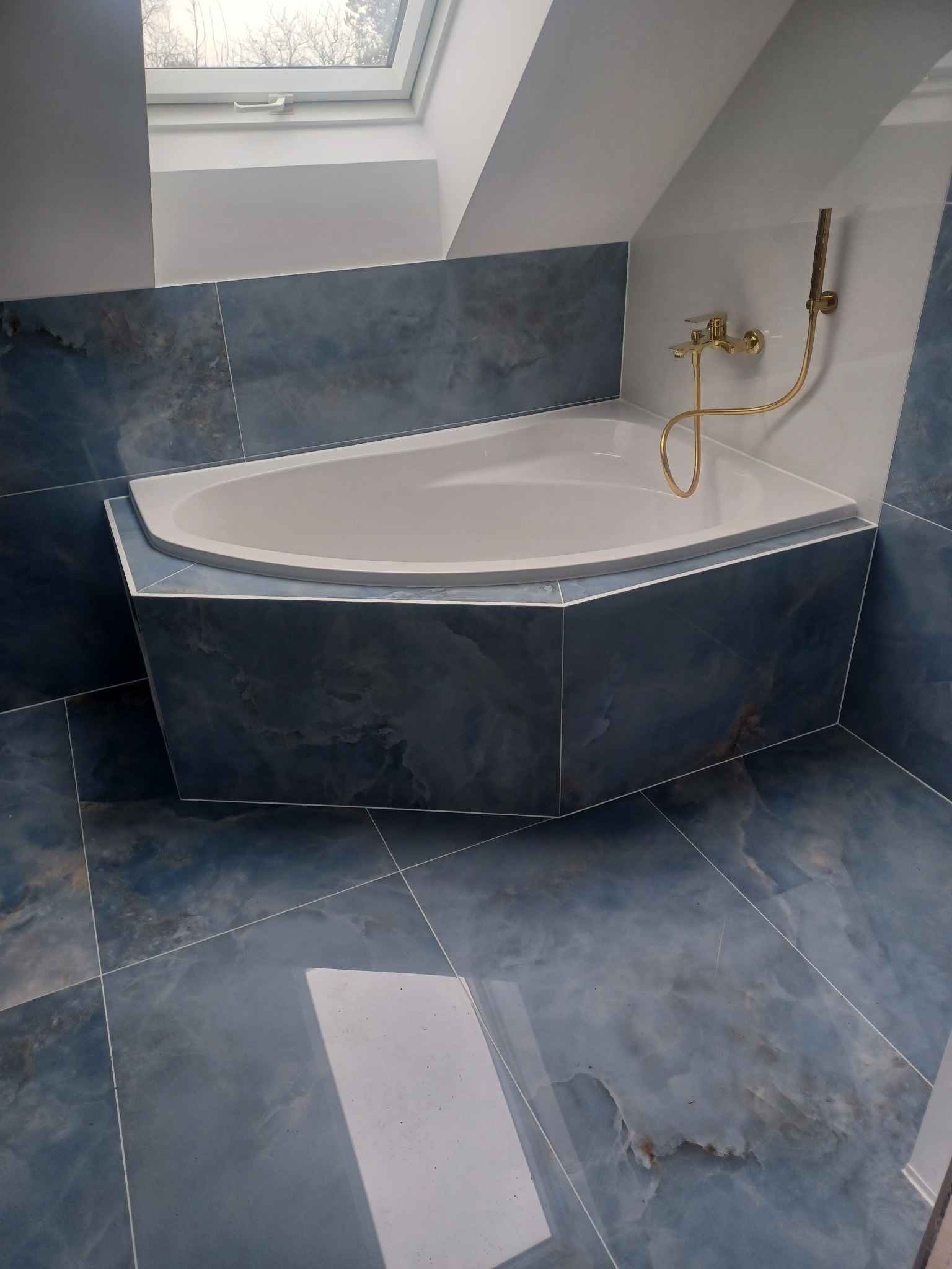 Narożna wanna obudowana płytkami imitującymi marmur w odcieniach błękitu i szarości, złota armatura, widok na okno dachowe, łazienka w Rzeszowie.