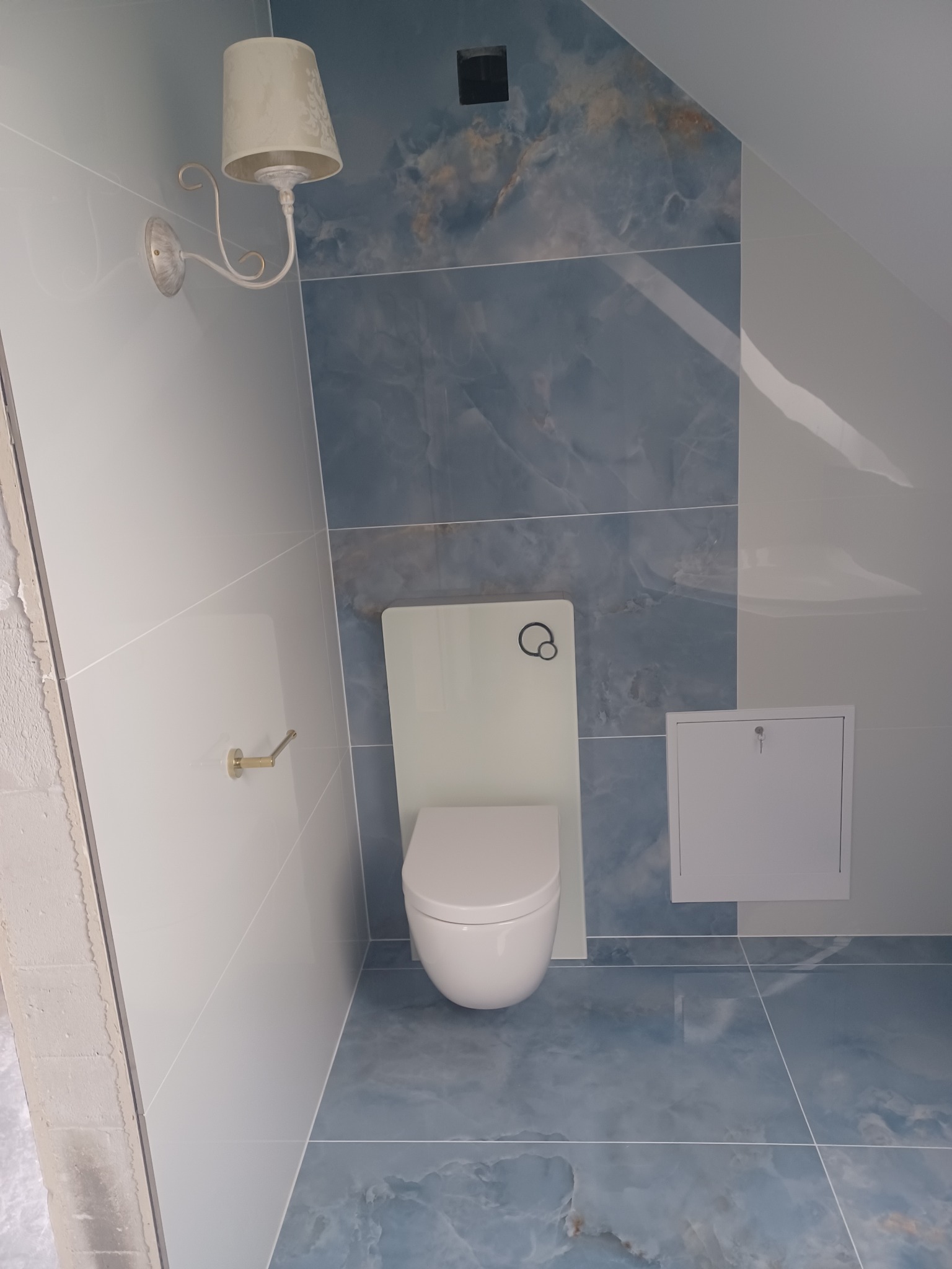 Wnętrze łazienki na poddaszu z białą wiszącą toaletą, niebieskimi płytkami imitującymi marmur, ozdobnym kinkietem i klapą rewizyjną.