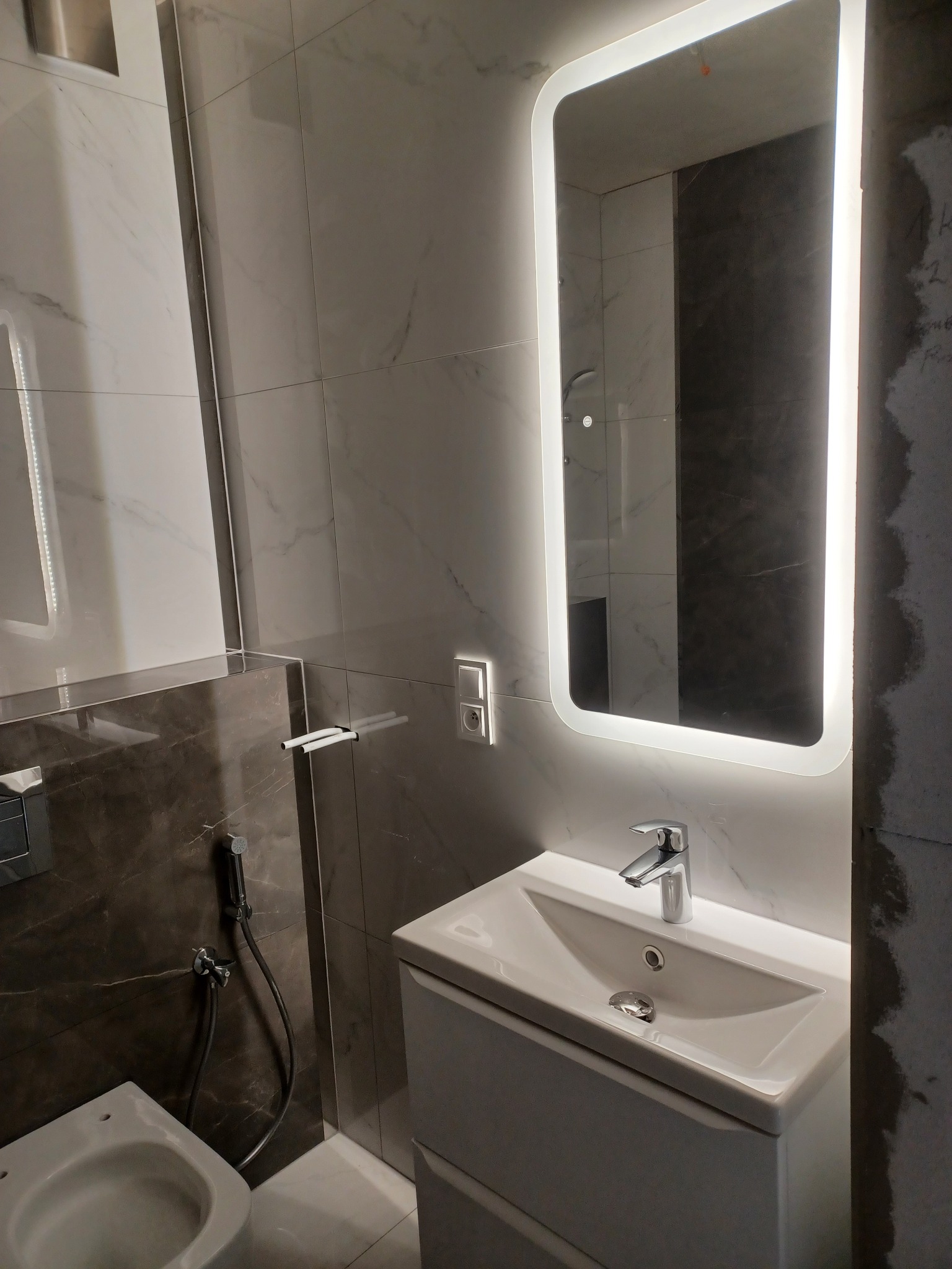 Nowoczesna łazienka z podświetlanym lustrem, umywalką z szafką i bidetką. Marmurowe płytki ścienne. Fragment toalety. Częściowo wykończone ściany.