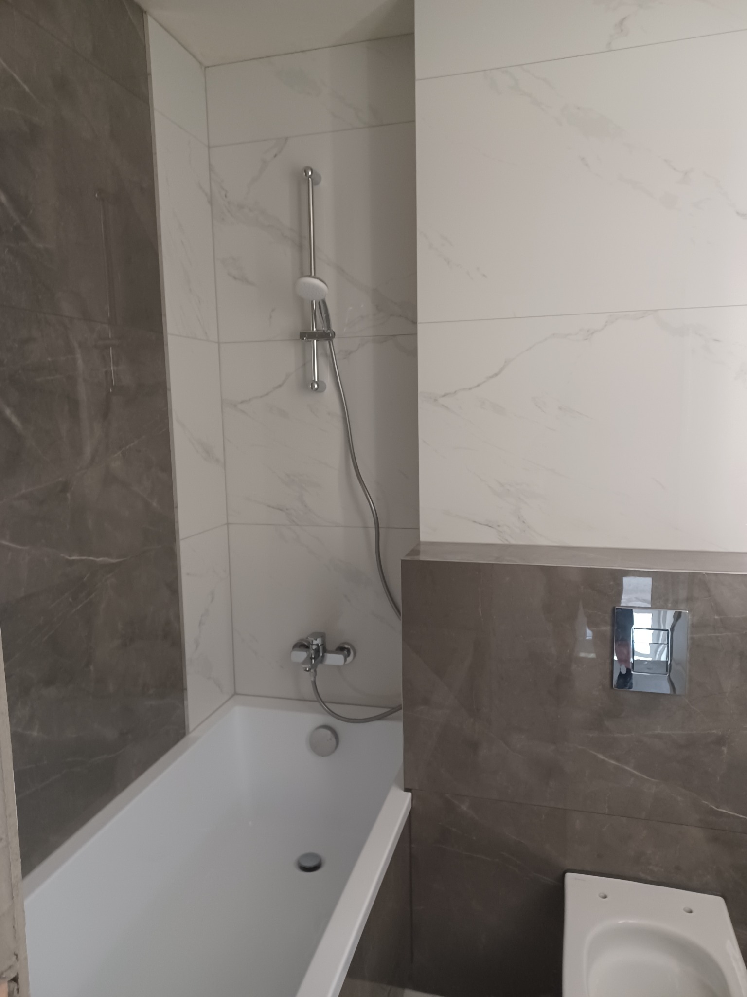 Nowoczesna łazienka w szaro-białej kolorystyce z wanną, prysznicem i podwieszaną toaletą. Widoczne marmurowe płytki na ścianach i chromowana armatura.