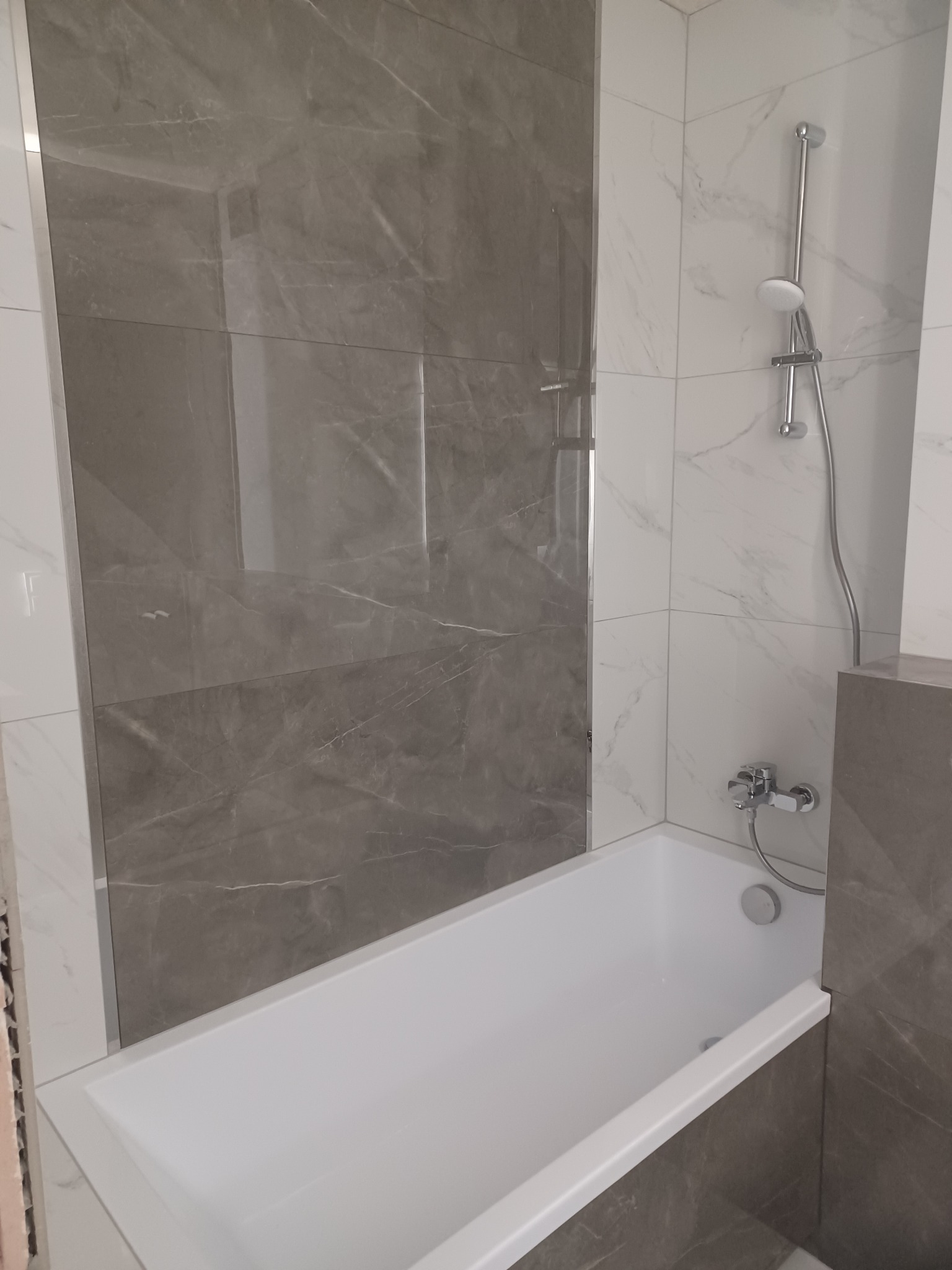 Biała wanna obudowana szarymi płytkami imitującymi marmur, z zamontowaną armaturą w Rzeszowie. Widoczny fragment ściany z szarymi i białymi płytkami.