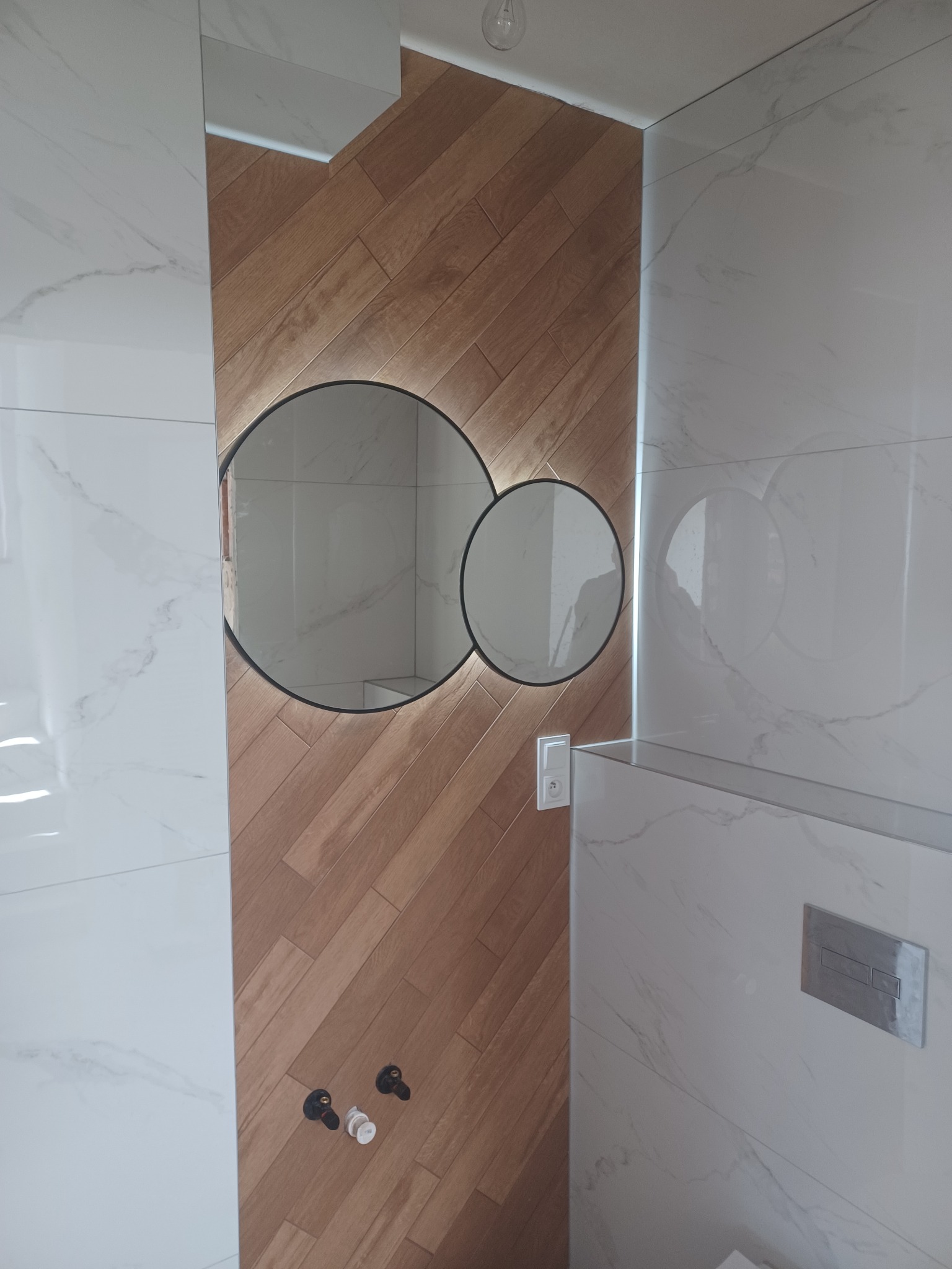 Nowoczesna łazienka: białe płytki marmurowe, drewnopodobna ściana z lustrem w kształcie okręgów, minimalistyczna armatura. Subtelne oświetlenie LED.