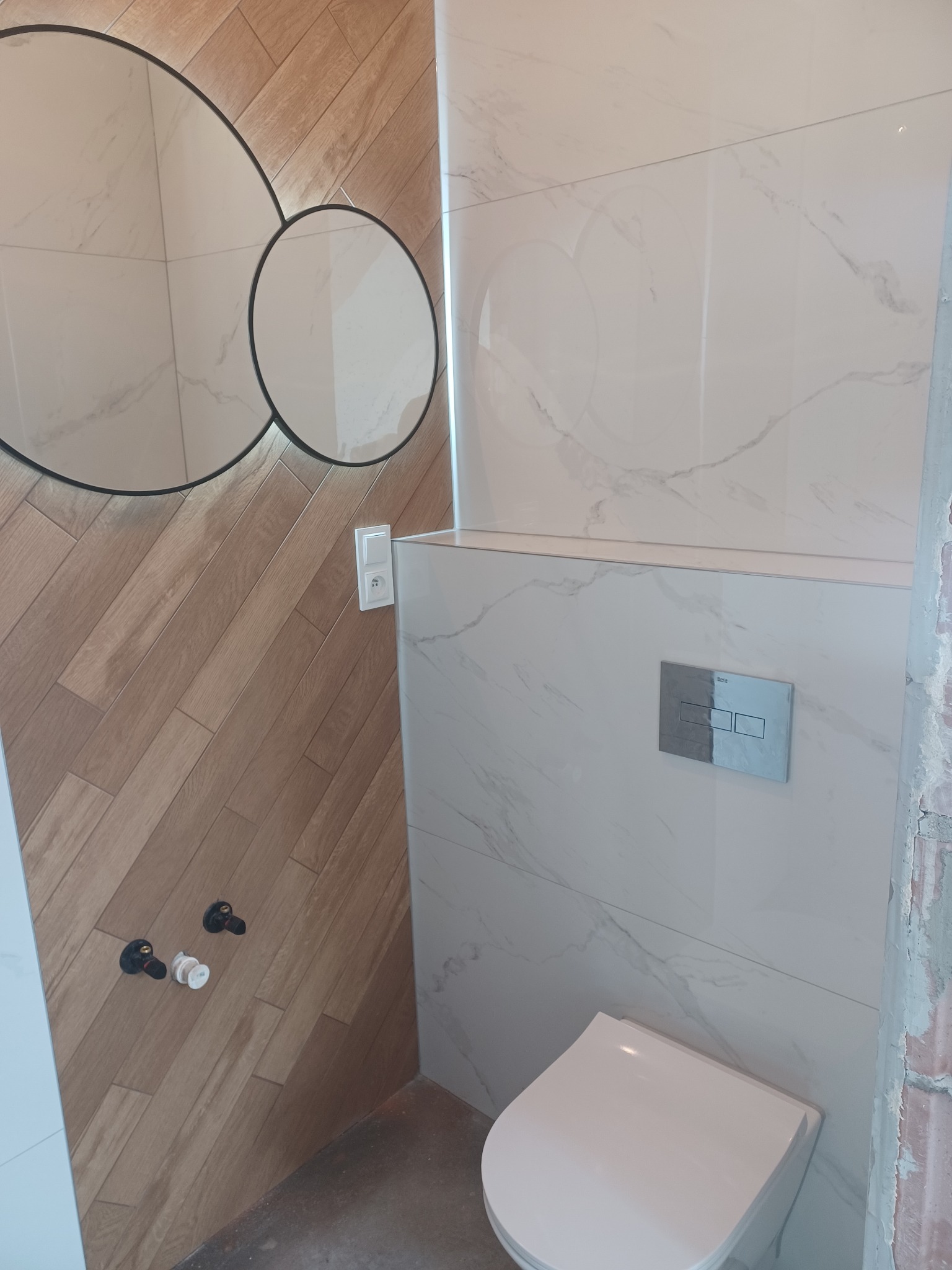 Nowoczesna toaleta w trakcie wykańczania: biała ceramika, marmurowe płytki i drewnopodobna ściana z okrągłym lustrem z podświetleniem. Widoczne elementy instalacji.
