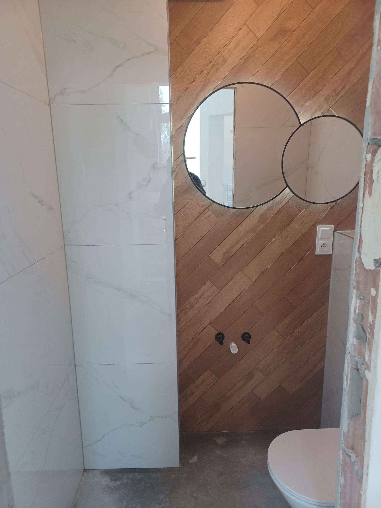 Wykończona łazienka w Rzeszowie z białymi płytkami imitującymi marmur i drewnopodobnymi ułożonymi w jodełkę. Na ścianie okrągłe lustro i biały sedes.