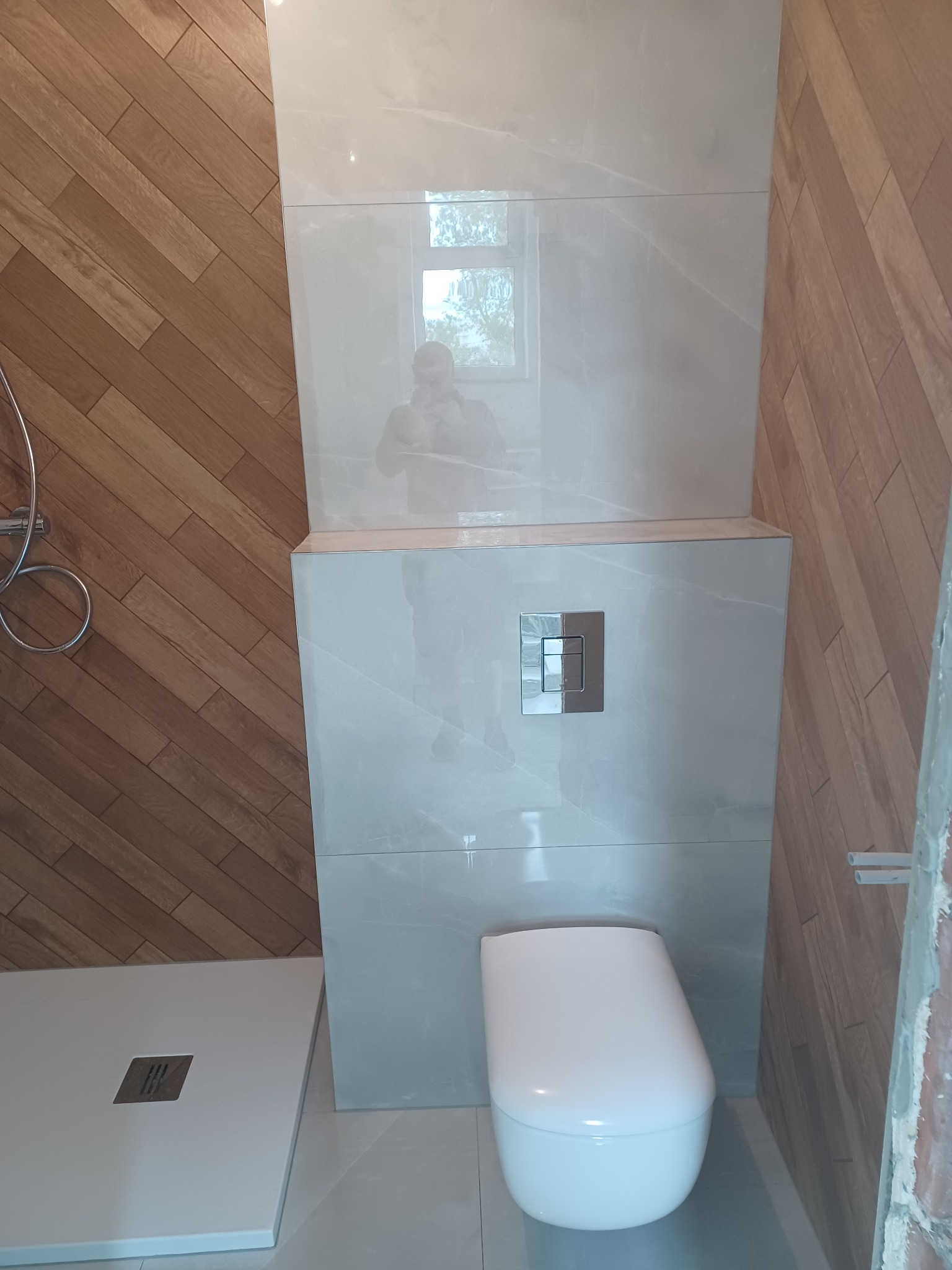 Wykończona łazienka z drewnopodobną okładziną i zabudową WC z płytkami imitującymi marmur, widoczny brodzik i fragment instalacji. Nowoczesny design.