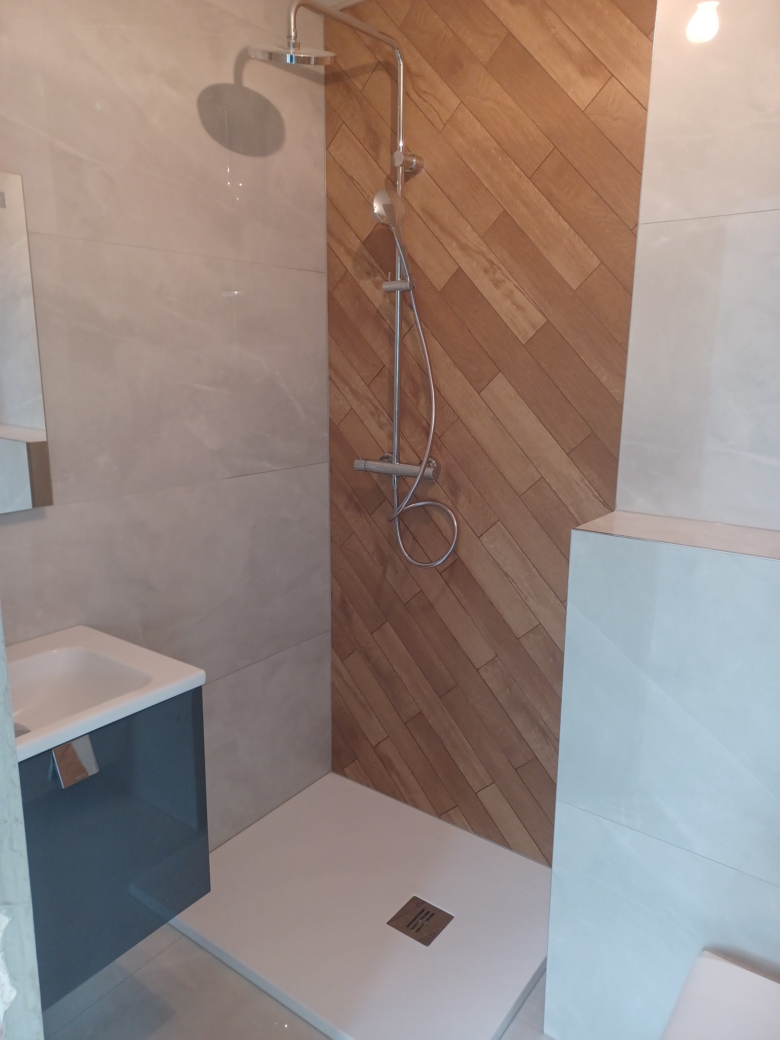 Nowoczesna łazienka z kabiną typu walk-in, wykończona płytkami imitującymi drewno na ścianie i szarymi płytkami na pozostałych ścianach. Umywalka z szafką w odcieniach szarości.