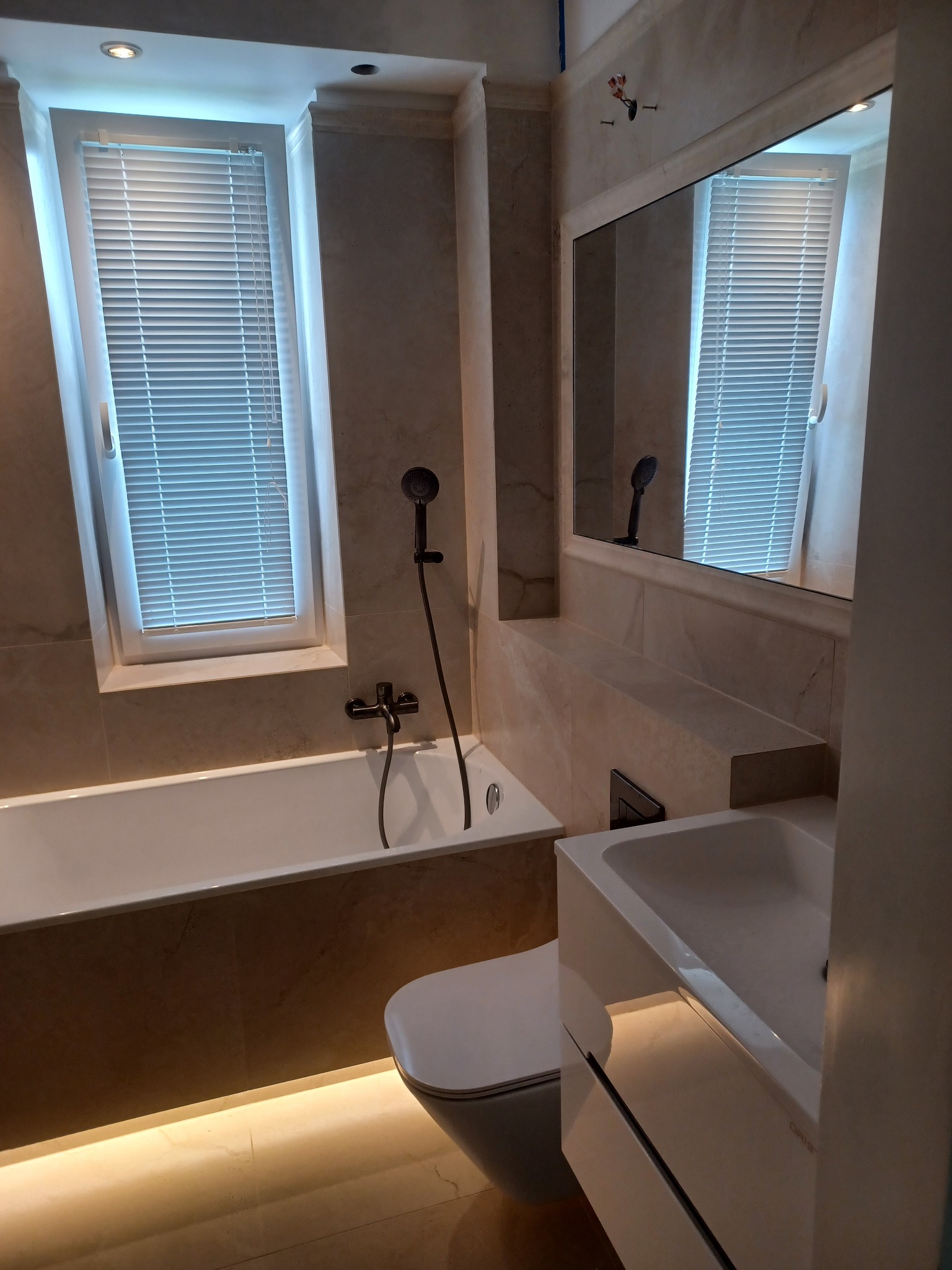 Elegancka łazienka w beżowych tonacjach z oknem, wanną, umywalką i toaletą. Podświetlenie LED podkreśla nowoczesny design. Widoczne marmurowe płytki.