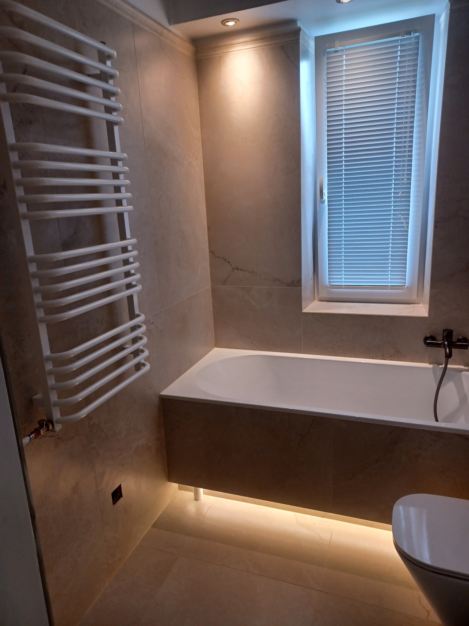 Wykończona łazienka w beżowych płytkach z podświetloną wanną, grzejnikiem drabinkowym i oknem z żaluzjami. Nowoczesny design i ciepłe oświetlenie.