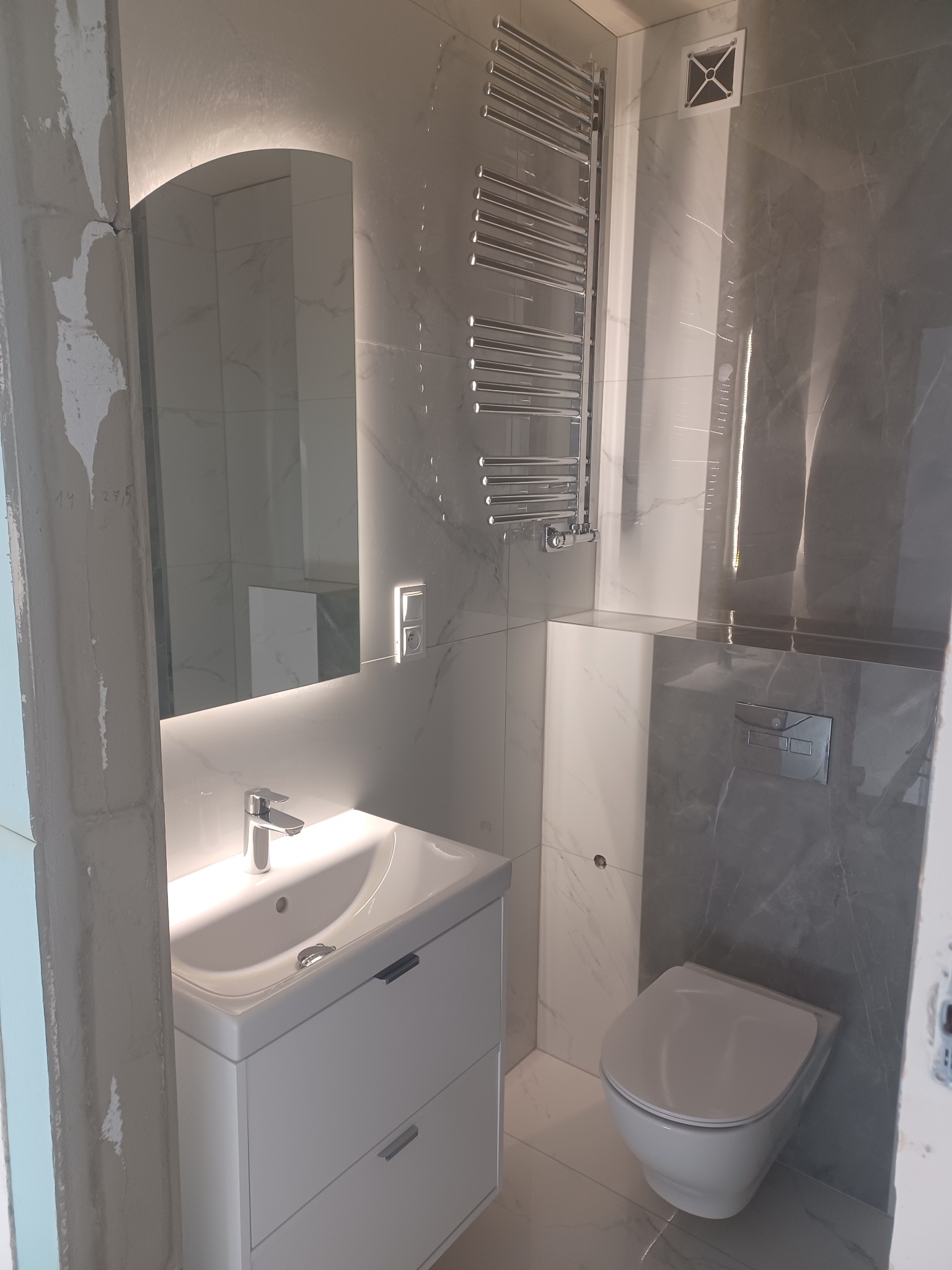 Nowoczesna łazienka w szaro-białych marmurkowych płytkach, z podświetlanym lustrem, grzejnikiem drabinkowym i podwieszaną toaletą. Elementy wykończenia w trakcie prac.