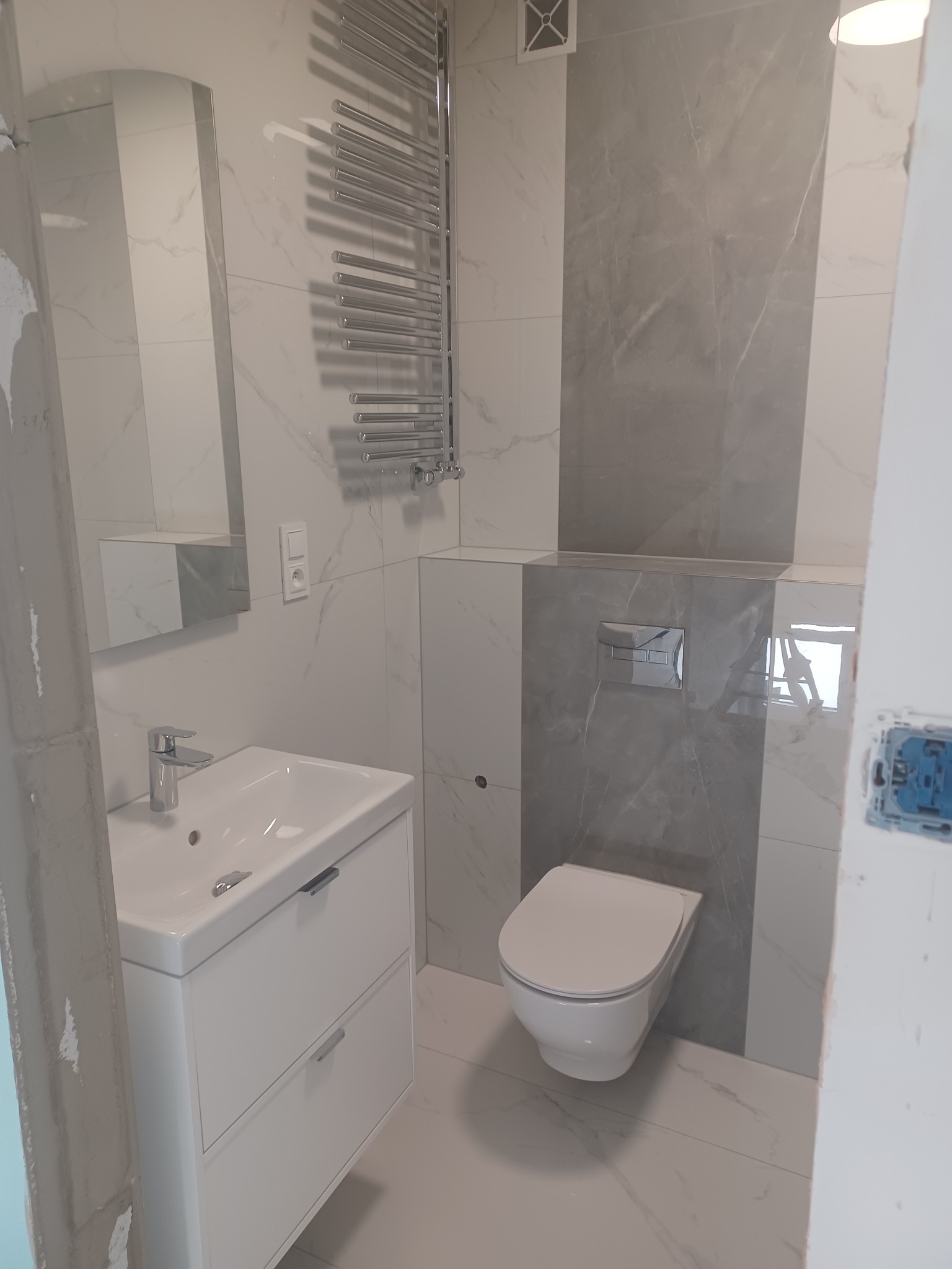 Wykończona łazienka z umywalką na szafce, toaletą i grzejnikiem drabinkowym na tle marmurowych płytek w odcieniach bieli i szarości. Nowoczesny design.