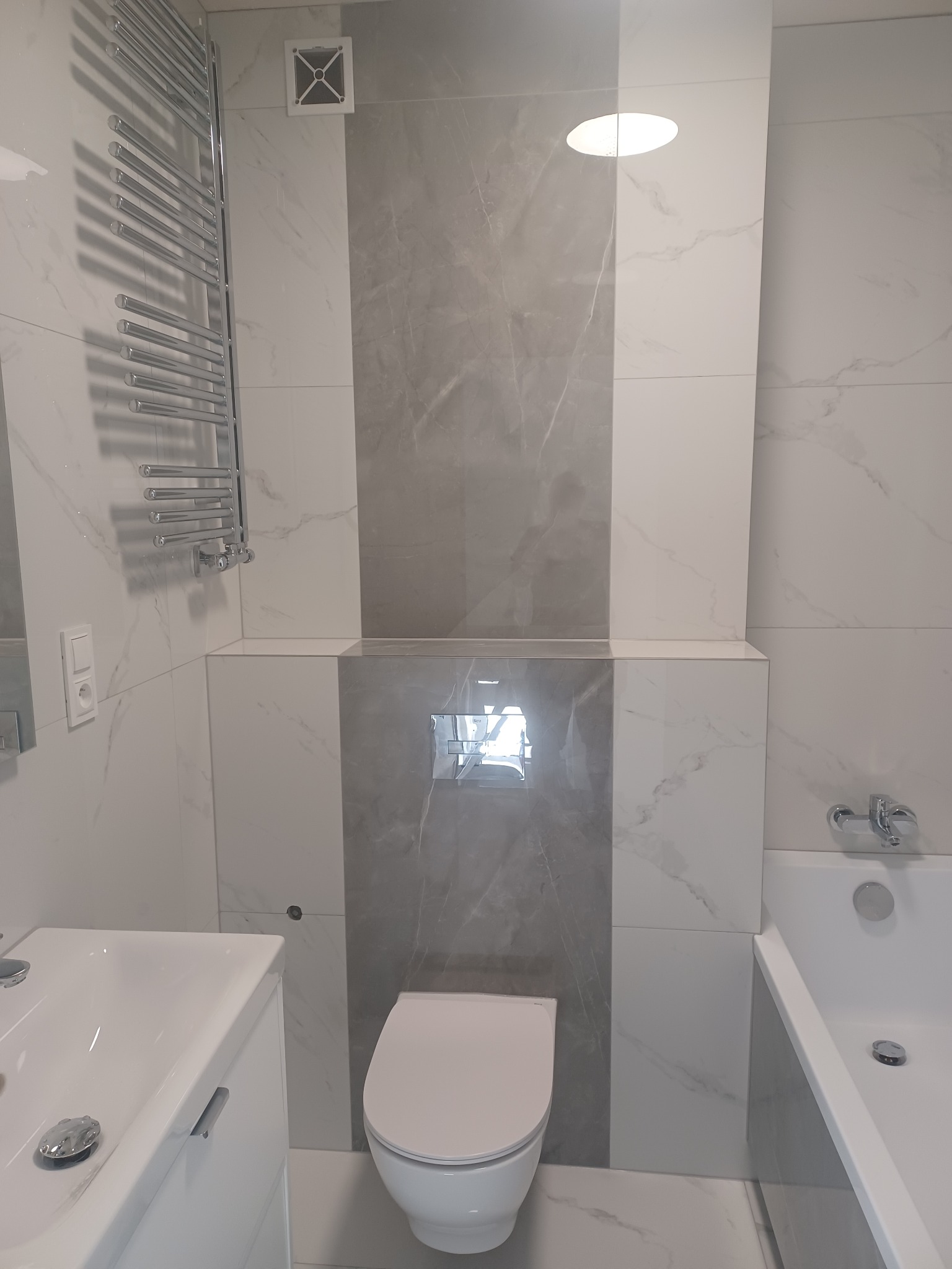 Nowoczesna łazienka z białymi i szarymi płytkami imitującymi marmur, wisząca toaleta, grzejnik drabinkowy i umywalka z szafką. Minimalistyczny design.